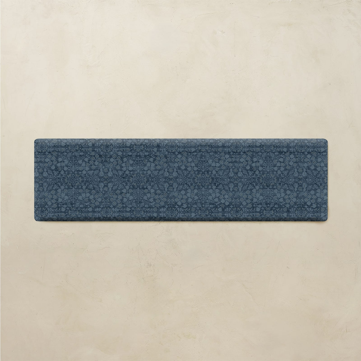 A rectangular blue textured runner rug with a subtle floral pattern, displayed horizontally against a warm beige textured wall background.