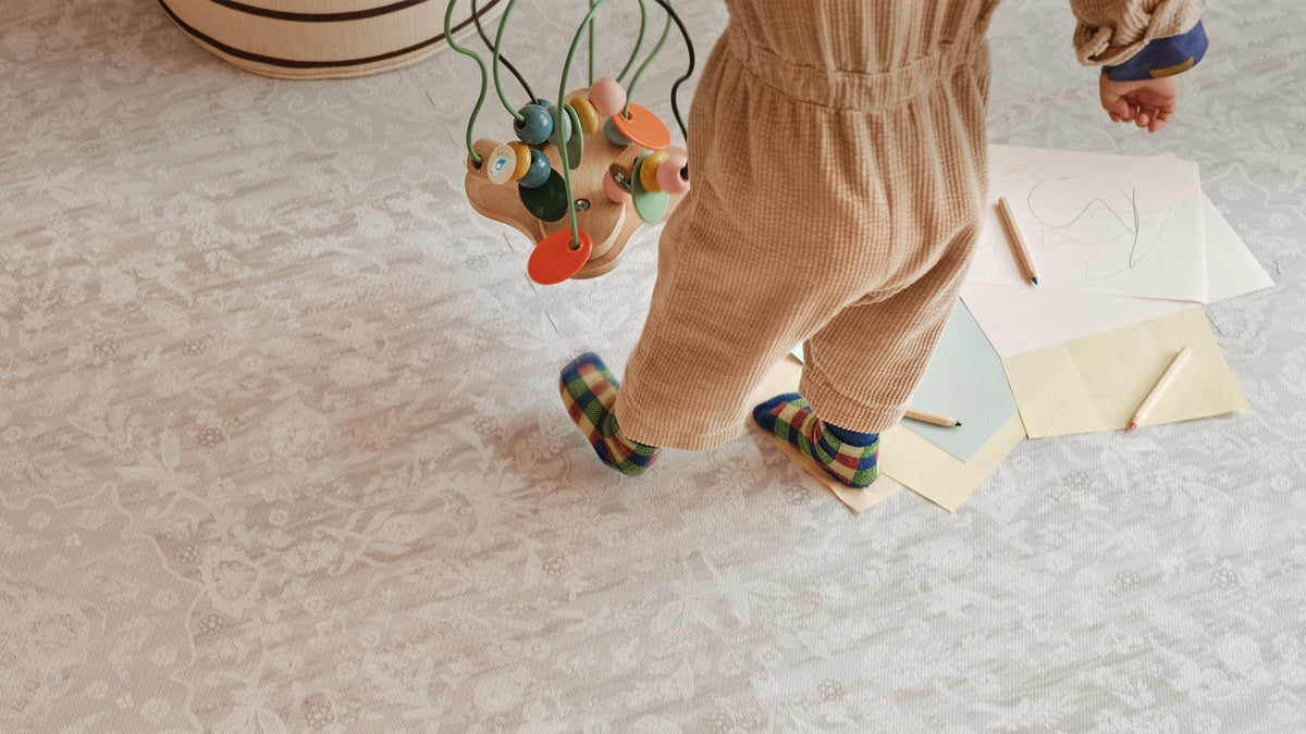 A child wearing beige corduroy pants and colorful plaid socks walks on a light patterned floor while holding a wooden bead toy. Papers and pencils are scattered nearby.