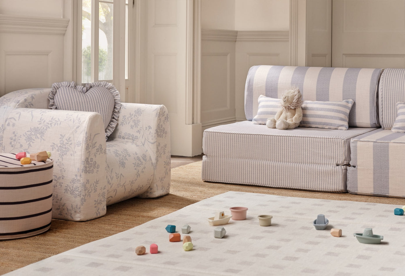 Children's toys on a tumbling mat in a play room with a striped play couch and floral play chair with ruffle pillows and an ottoman.