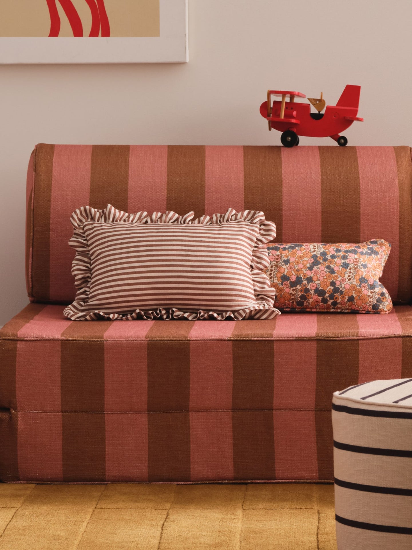 A striped upholstered sofa in pink and brown tones with two decorative pillows, a small red toy airplane on the backrest, and part of a striped ottoman in the foreground.