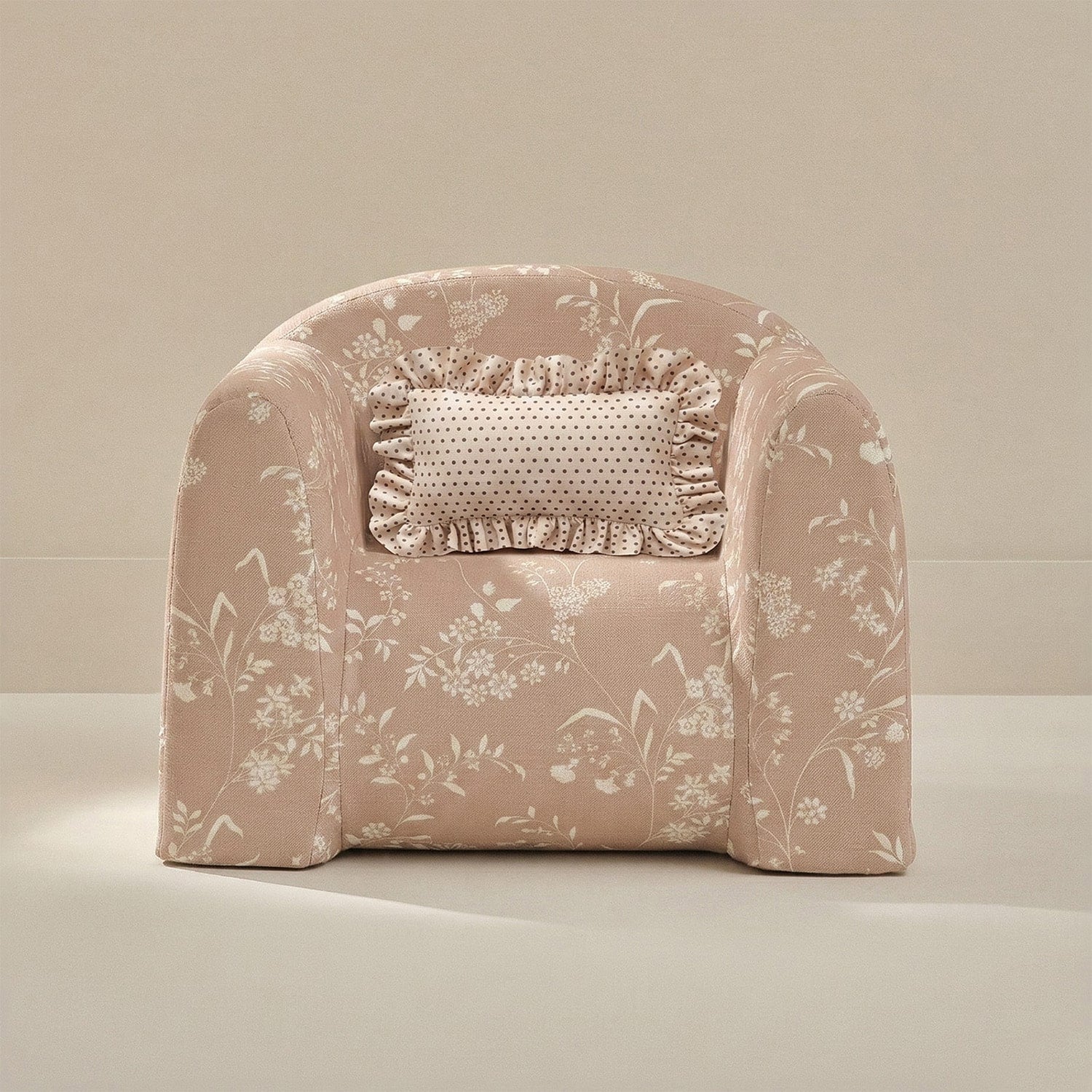A small upholstered armchair in a dusty blush pink floral fabric, featuring a matching ruffled accent pillow, set against a warm beige studio background.