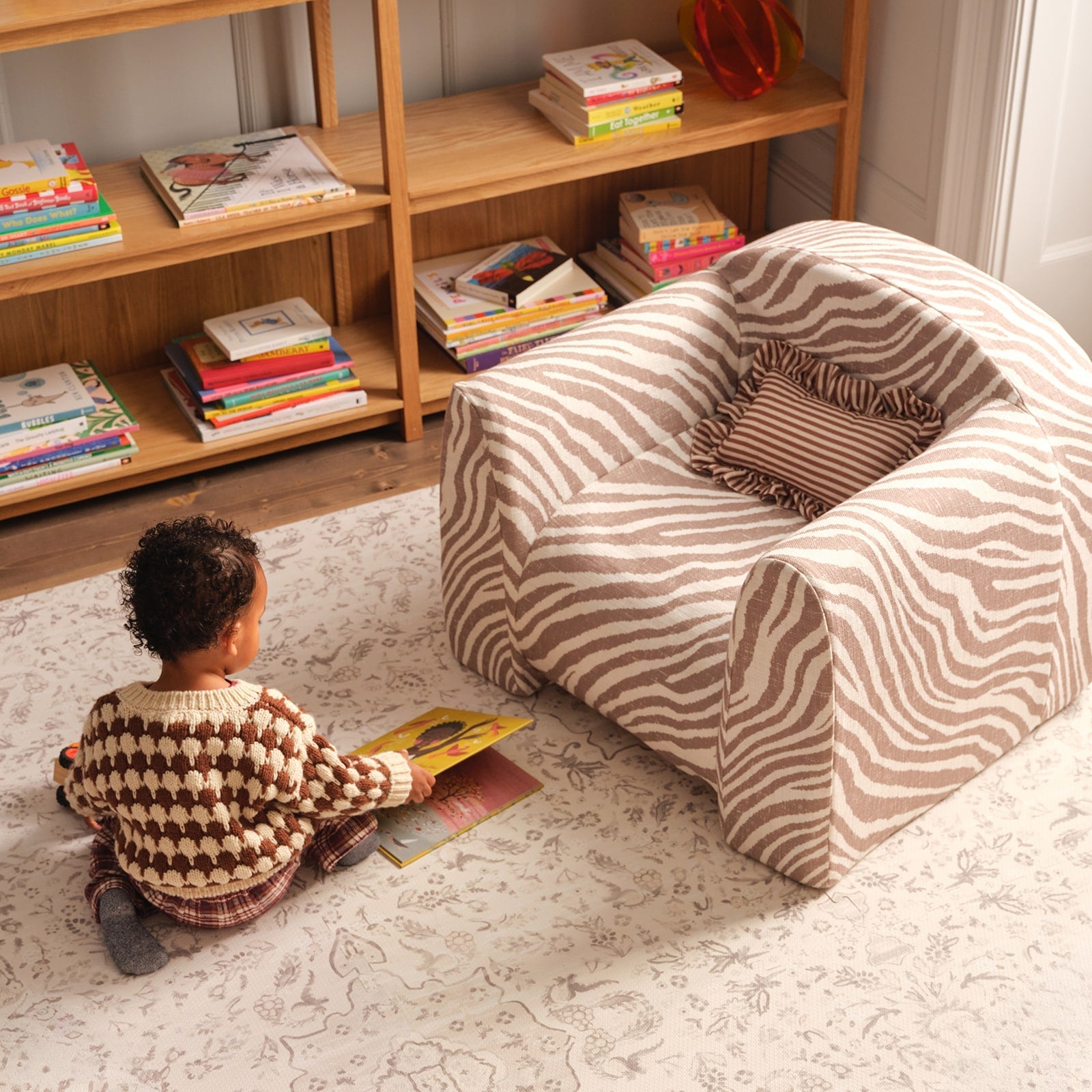 Child reading a book on a patterned play mat next to an animal striped play chair with a ruffle pillow