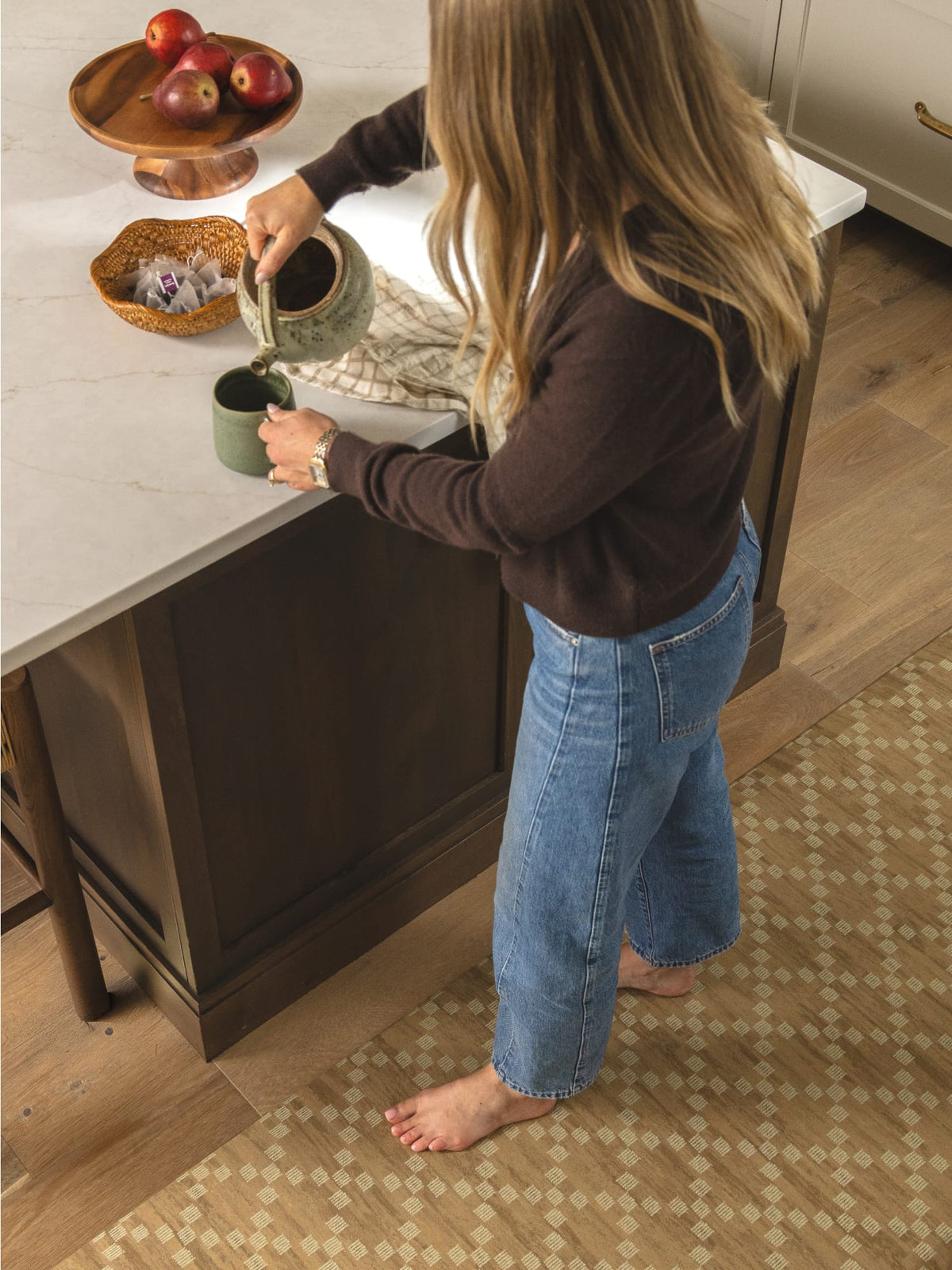 woman standing at the kitchen counter pouring herself a cup of tea while standing on a brown geometric pattern rug