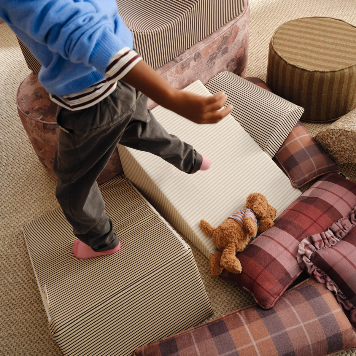 A child in pink socks steps across striped foam wedges and blocks, with a plush teddy bear resting on a cream striped wedge.