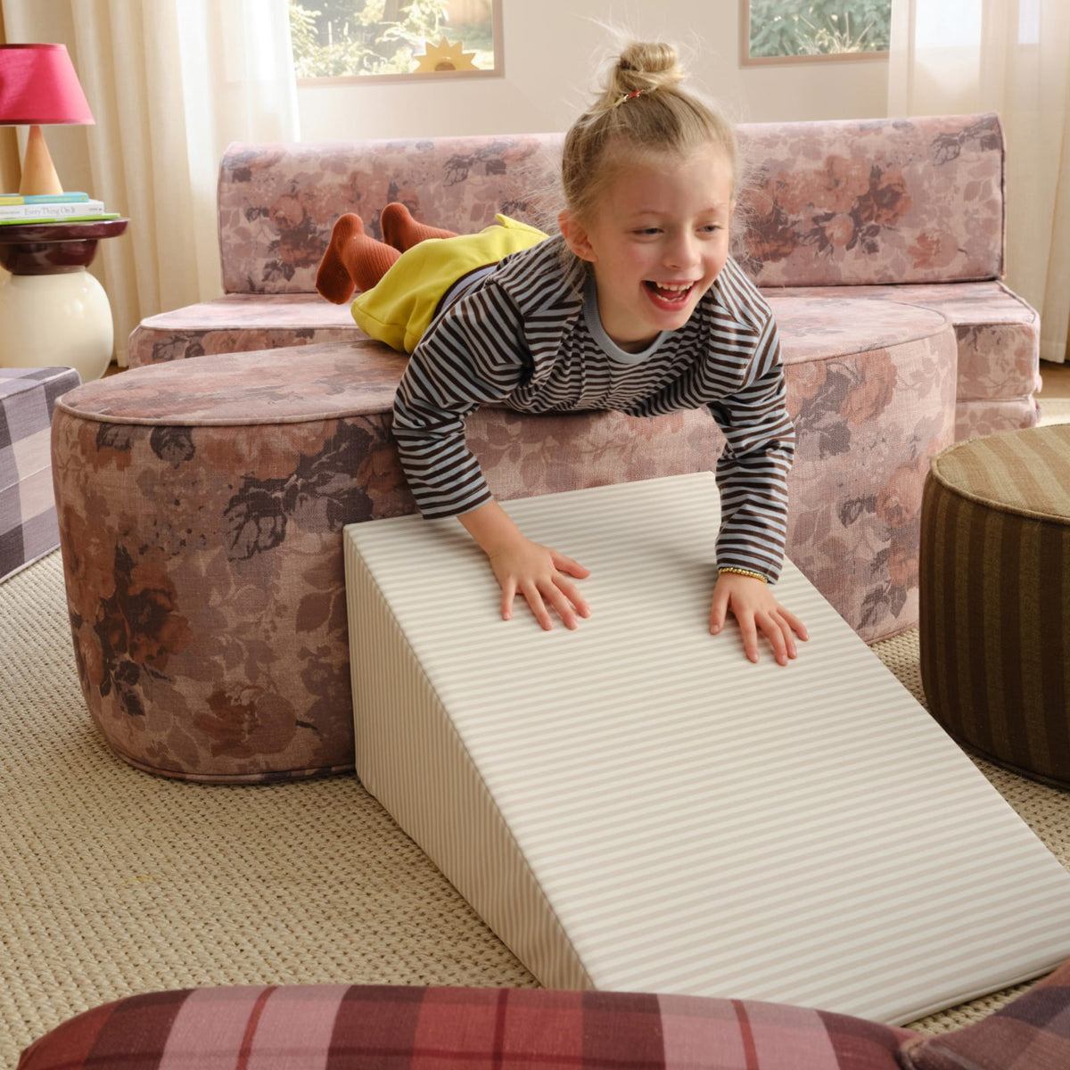 A laughing girl climbs down a cream striped wedge from a floral modular play couch.