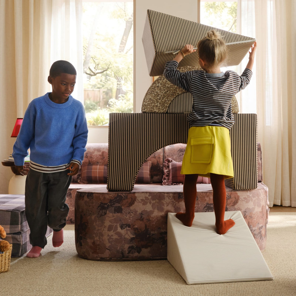 A girl stands on a cream striped wedge to stack a striped block on top of an arch while a boy in a blue sweater looks on.