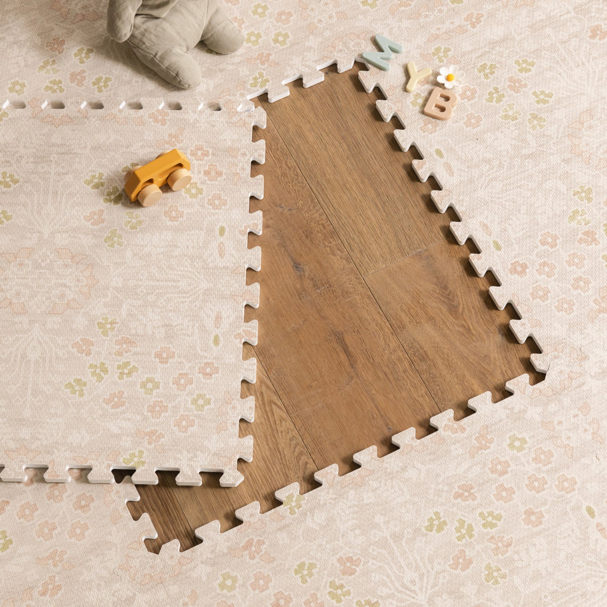 A corner of a blush floral foam play mat lifted back to show the wood floor beneath, with a baby's feet, a wooden toy car, and alphabet blocks nearby.