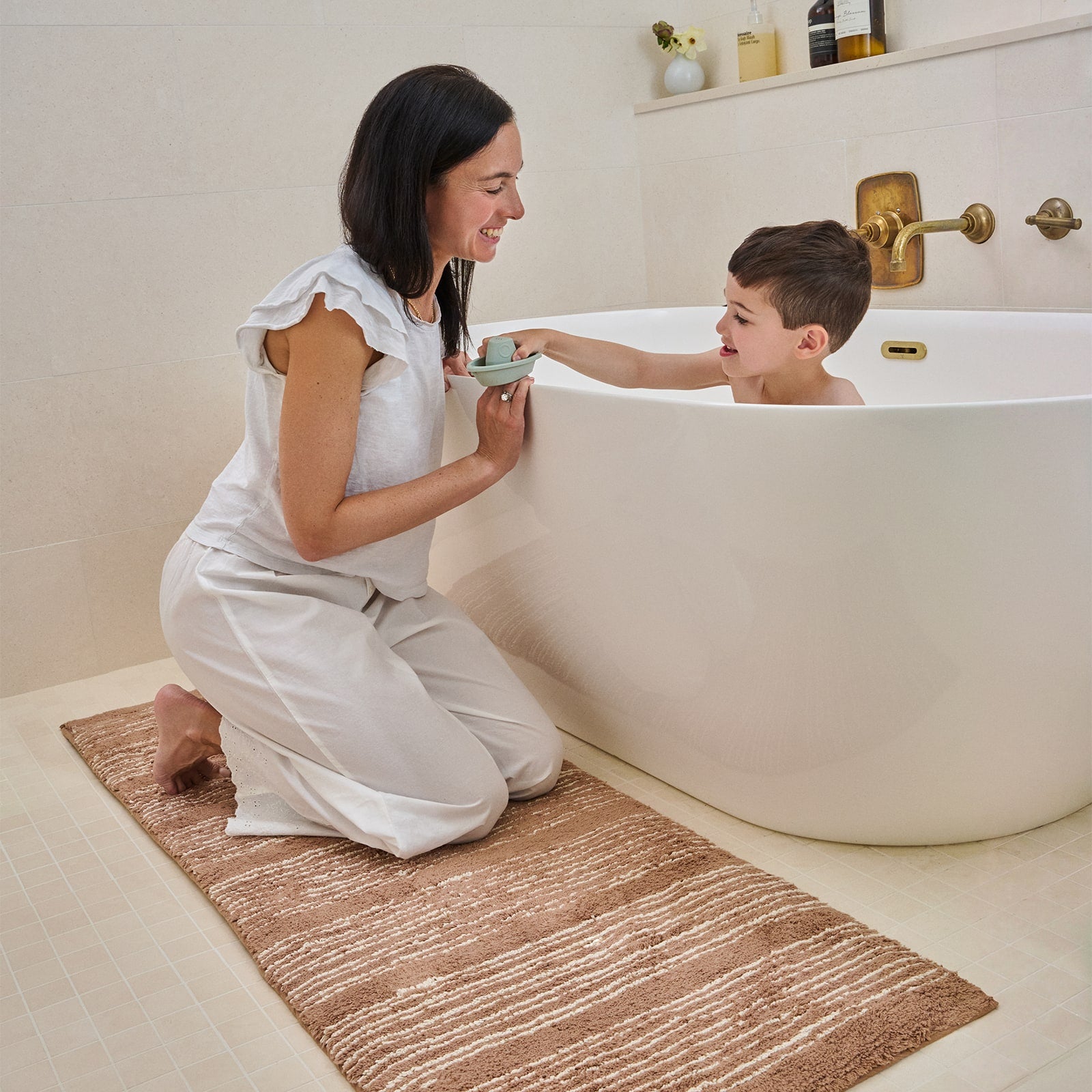 Bathroom with mom kneeling on a brown striped bath mat while son plays in the tub.