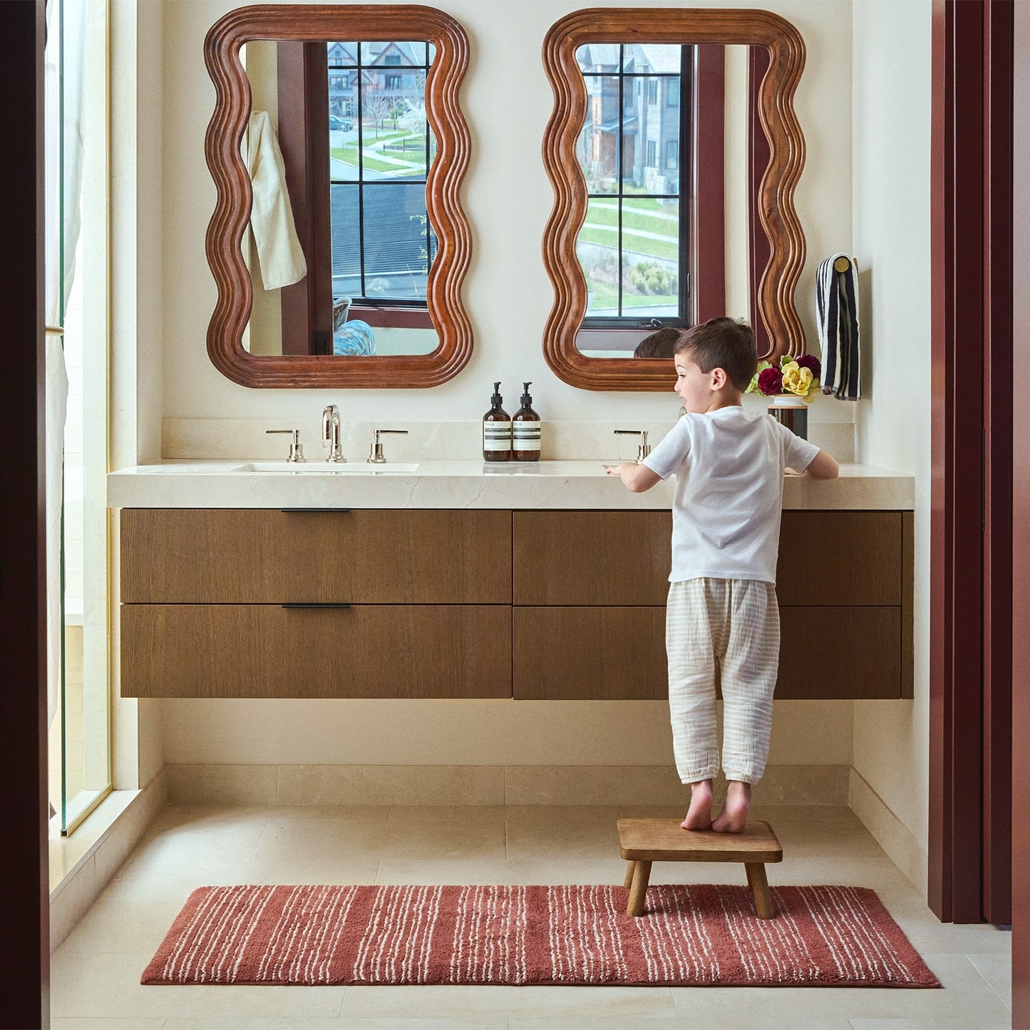 Child standing on a step stool in a modern bathroom with red and white stripe bath rug and wooden vanities and mirrors.