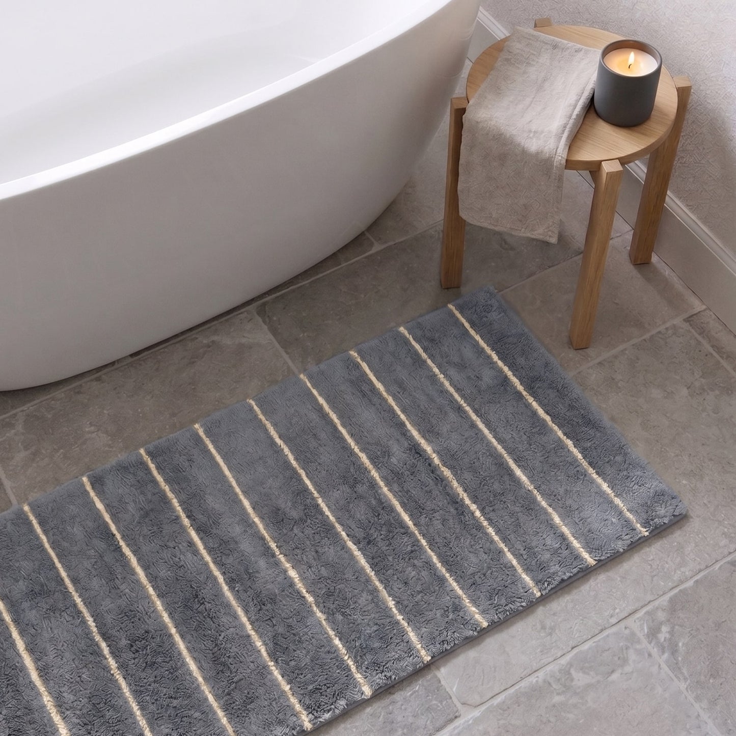 Gray bath mat with white stripes on a bathroom floor, next to a bathtub and stool with a candle.