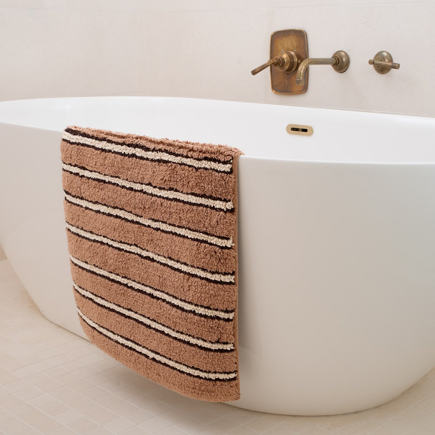 Brown striped bath mat hanging over a white bathtub with bronze faucet.