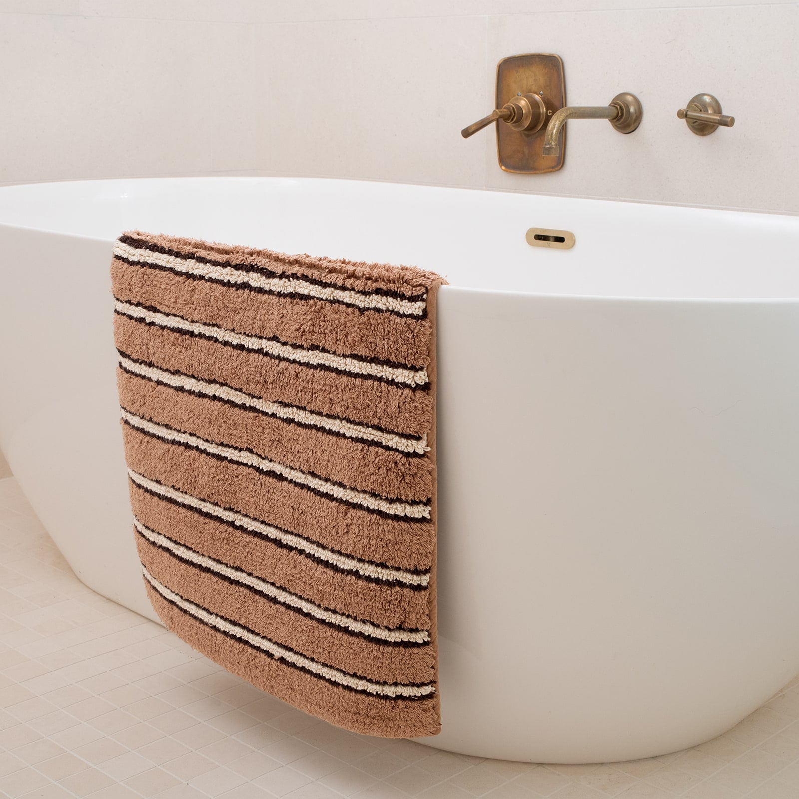 Brown striped bath mat hanging over a white bathtub with bronze faucet.