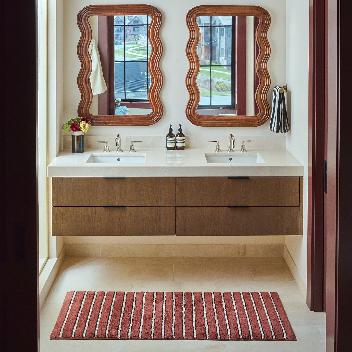 Red striped bath mat in front of double sinks in a bathroom with wooden mirrors and vanity.