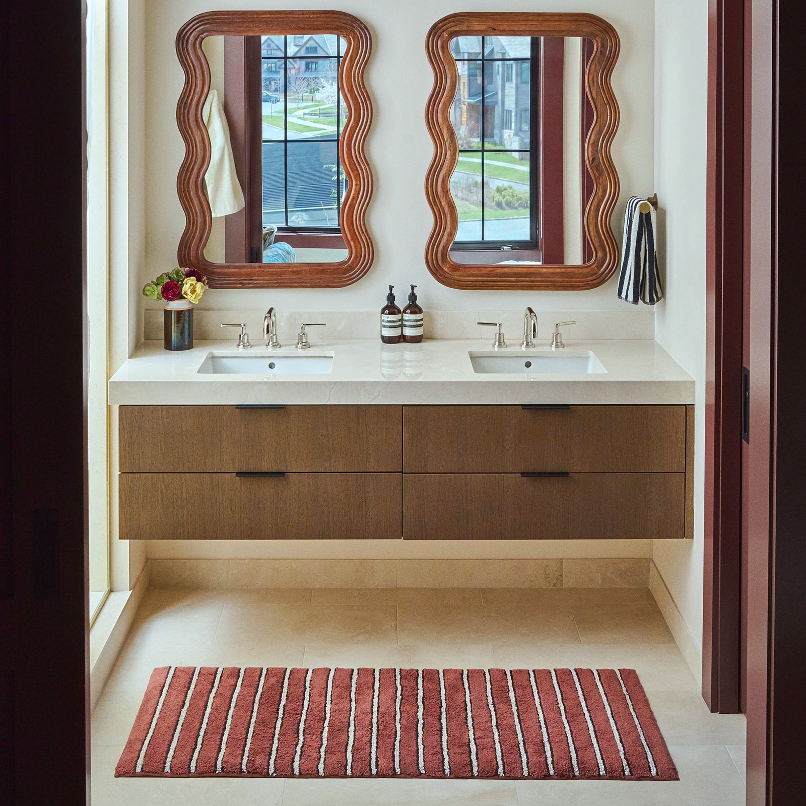 Red striped bath mat in front of double sinks in a bathroom with wooden mirrors and vanity.