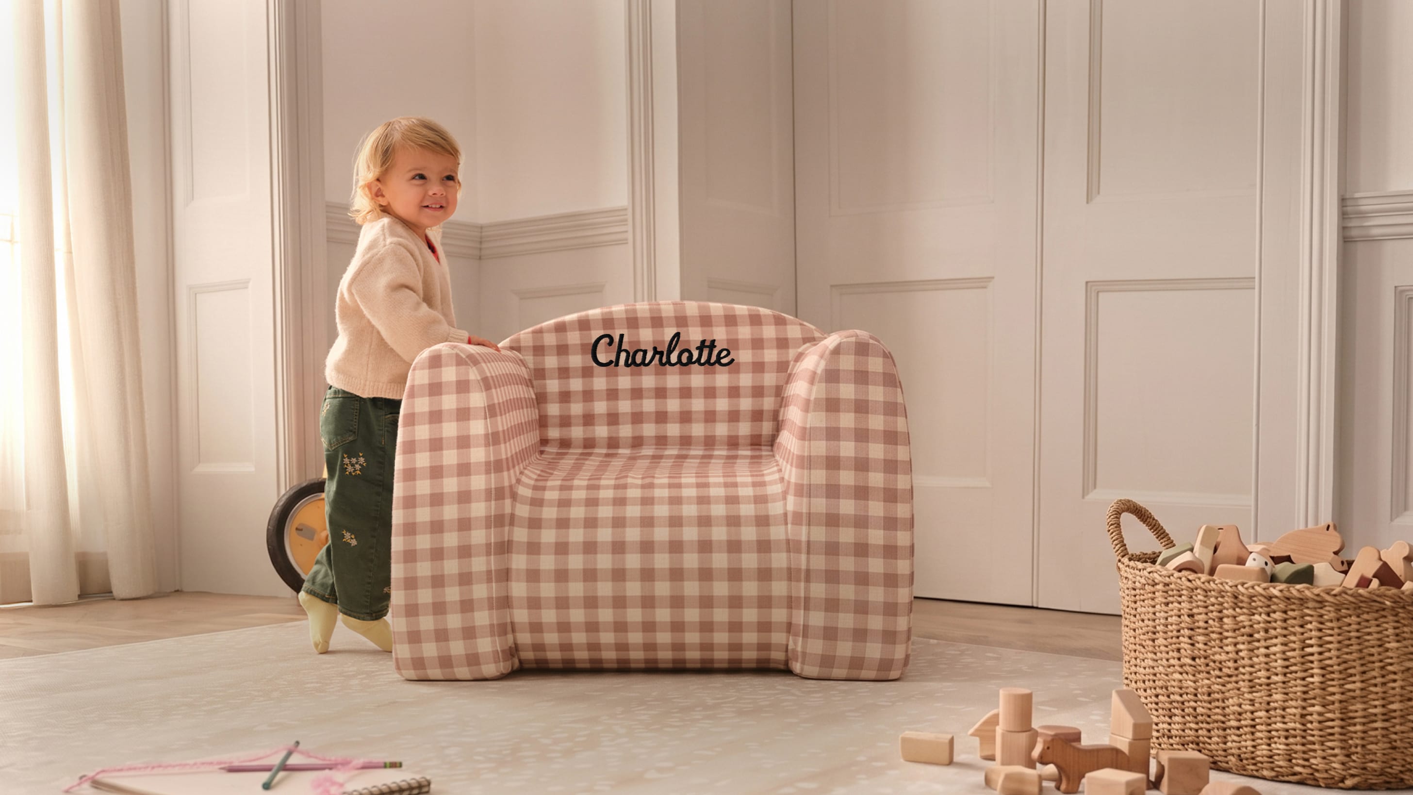 Child standing next to a checkered chair with 'Charlotte' on it in a room with toys and a basket.