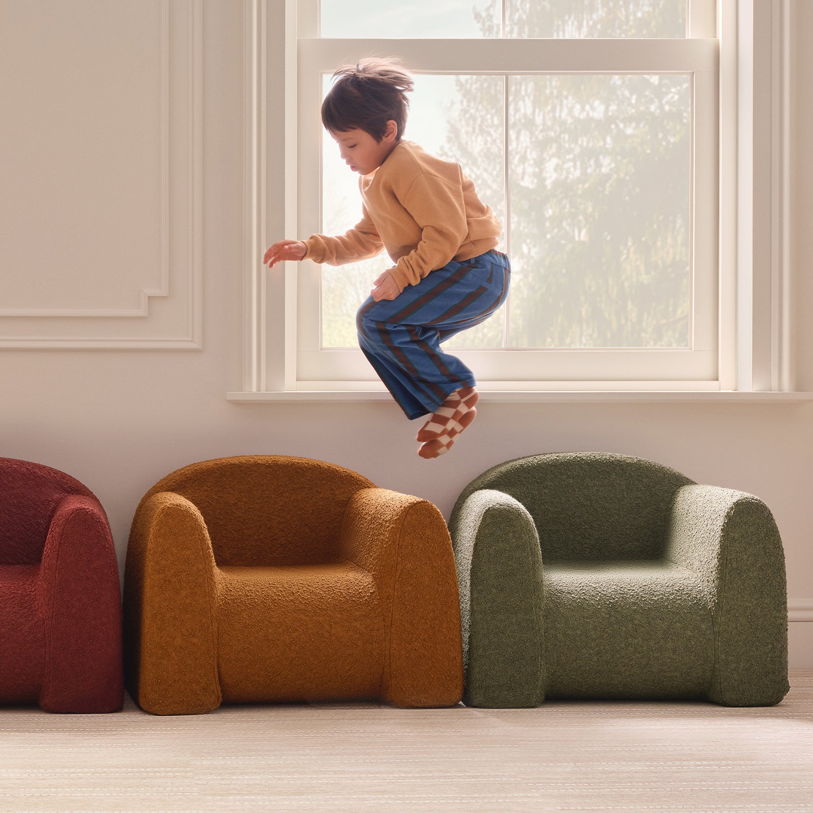 Child jumping on a colorful armchair in a room with large windows.