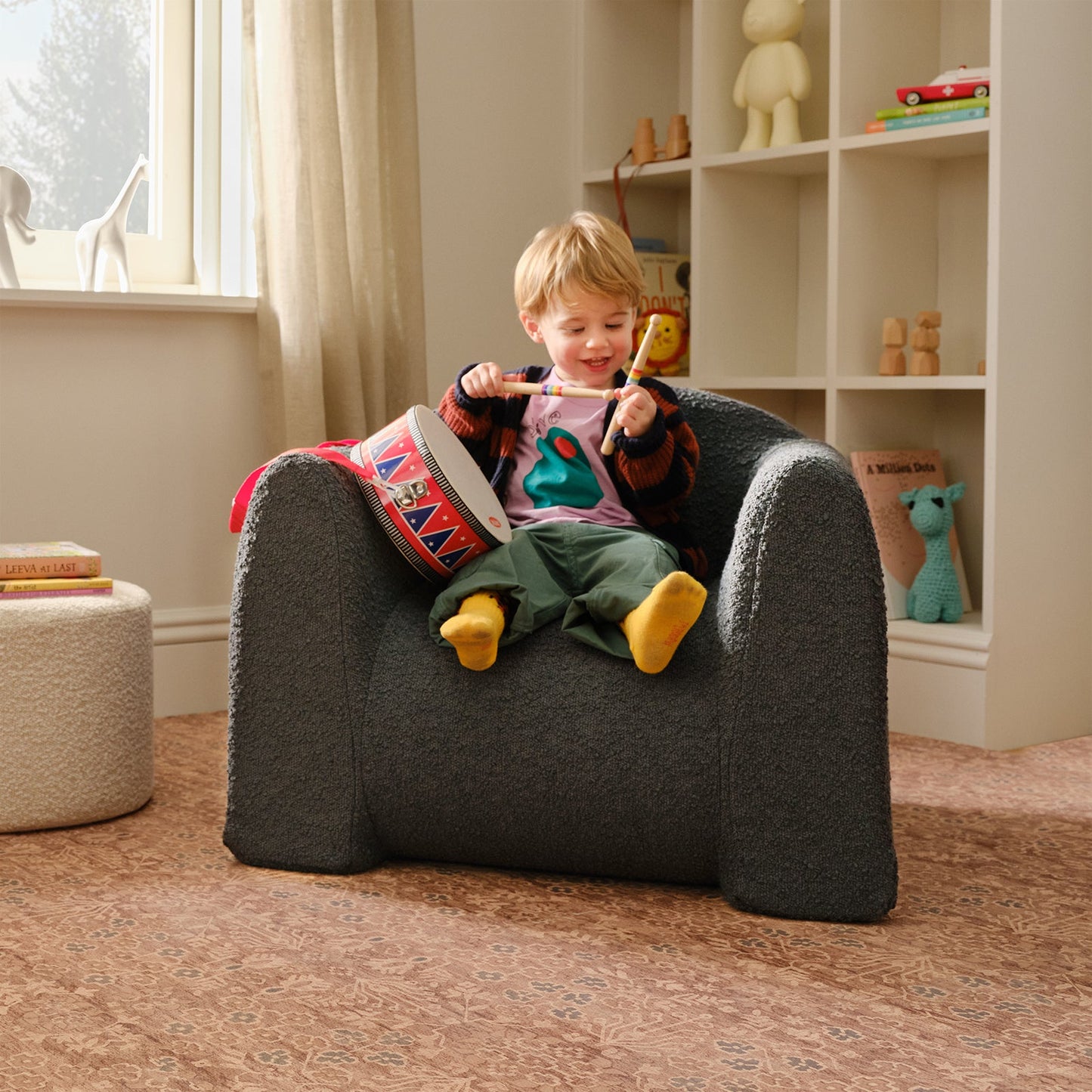 Child sitting on a gray chair in a room with books and toys.