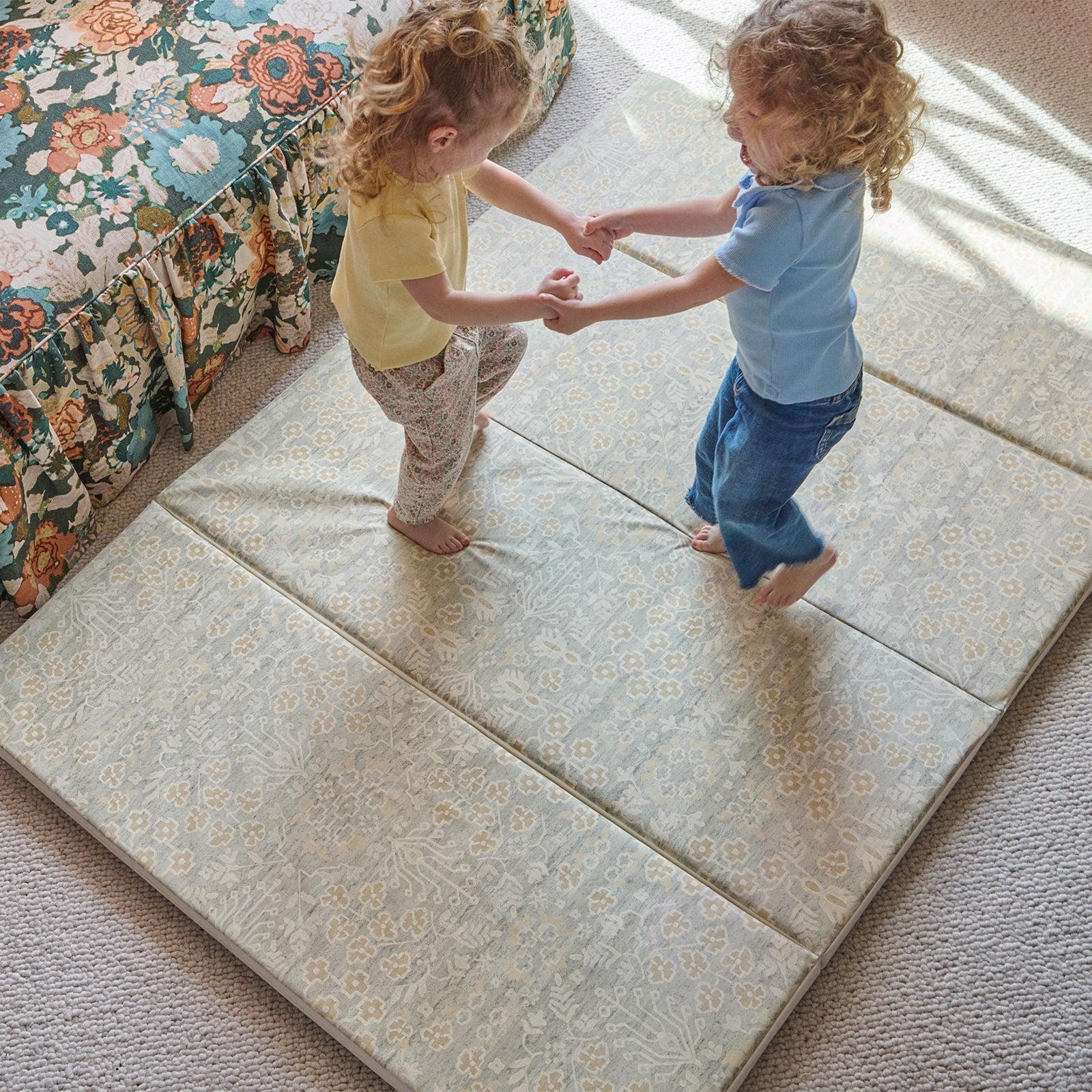 Two children holding hands on a patterned tumbling mat in a room with a floral pattern couch.