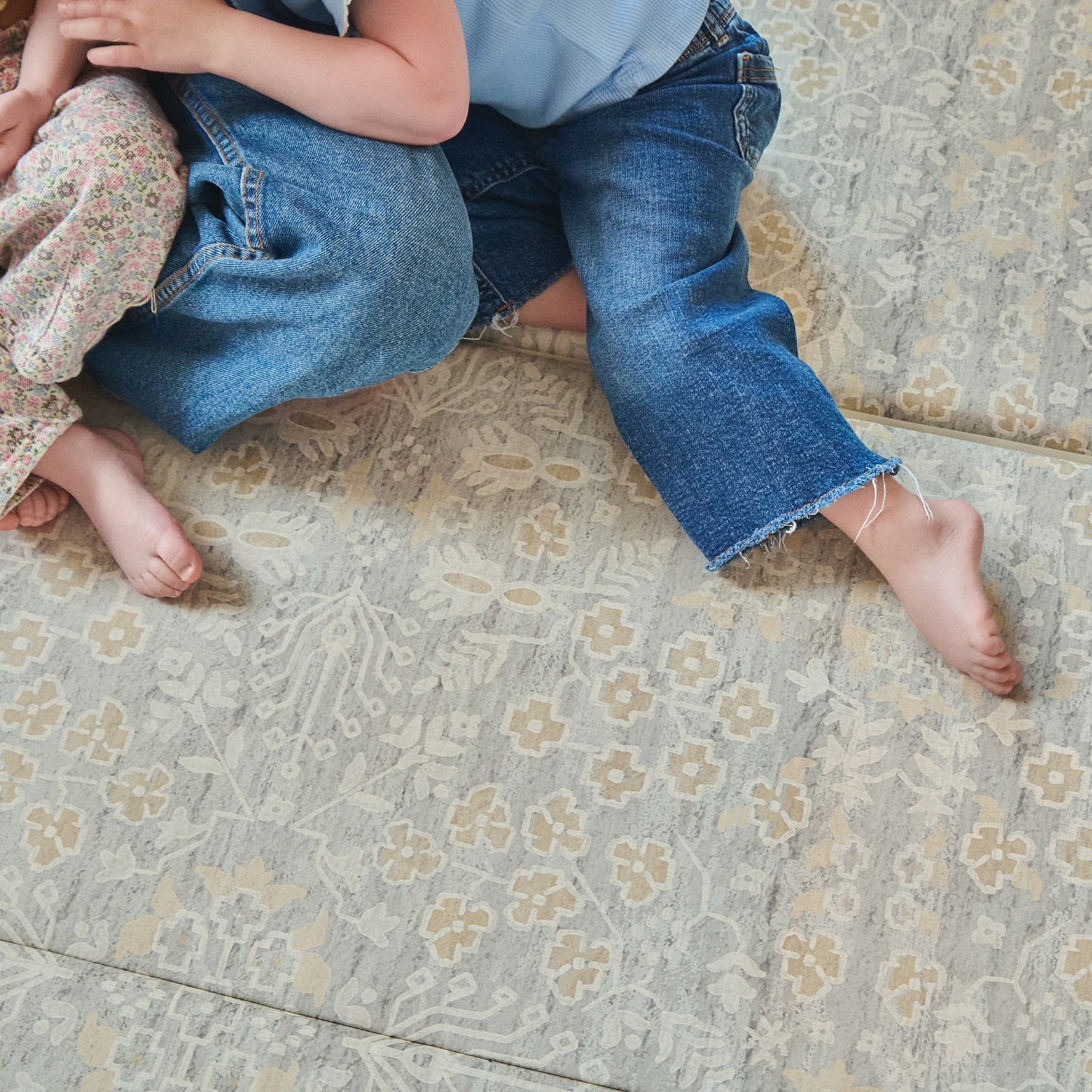 Two children sitting on a patterned tumbling mat with floral pants and blue jeans.