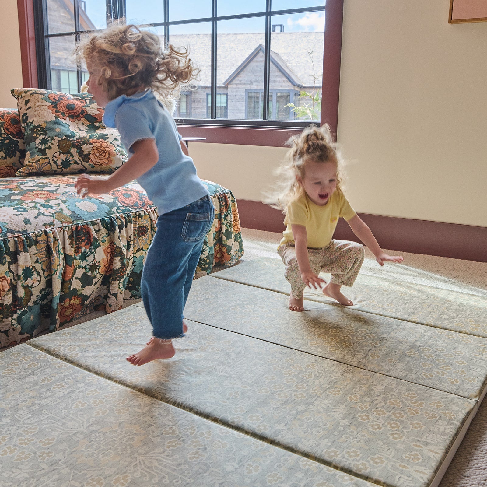 Two children playing on a large tumbling mat in a room with a couch and window.