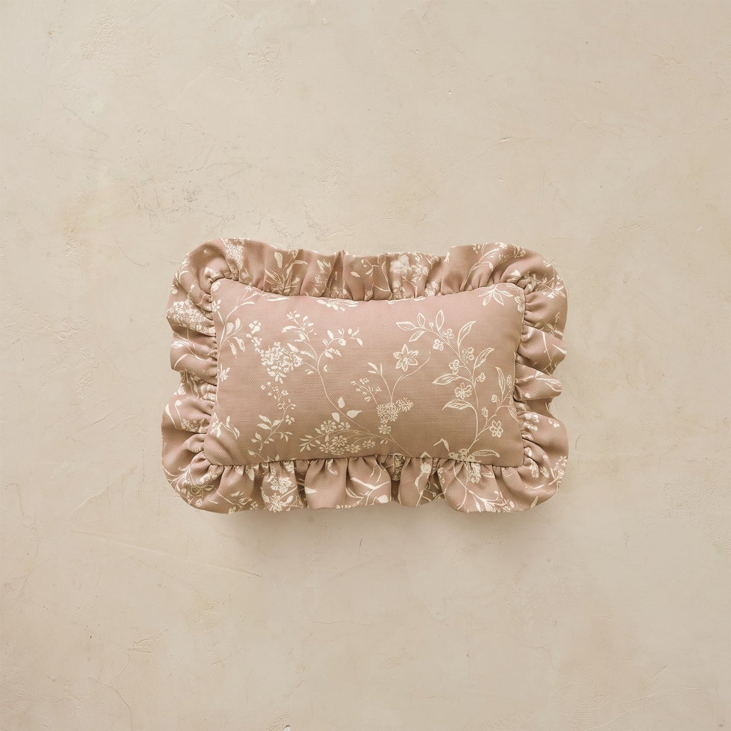 A rectangular decorative pillow with ruffled edges and a subtle floral pattern, shown against a textured beige background.