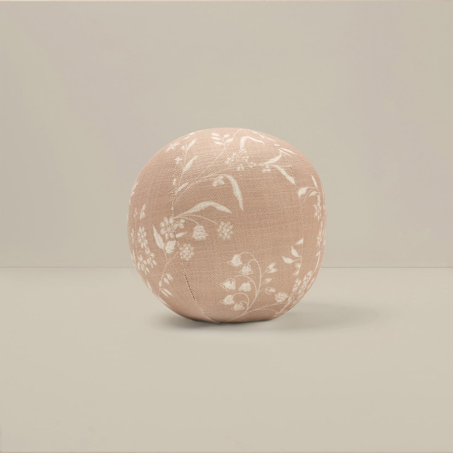 A round, upholstered cushion or pouf in a dusty blush pink fabric with a white floral pattern, set against a neutral beige background.