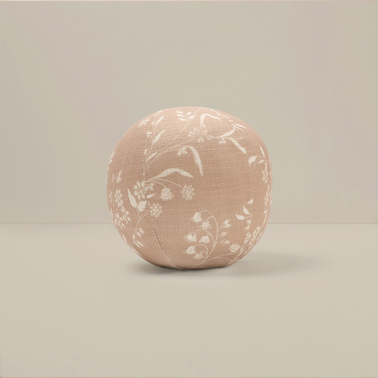 A round, upholstered cushion or pouf in a dusty blush pink fabric with a white floral pattern, set against a neutral beige background.