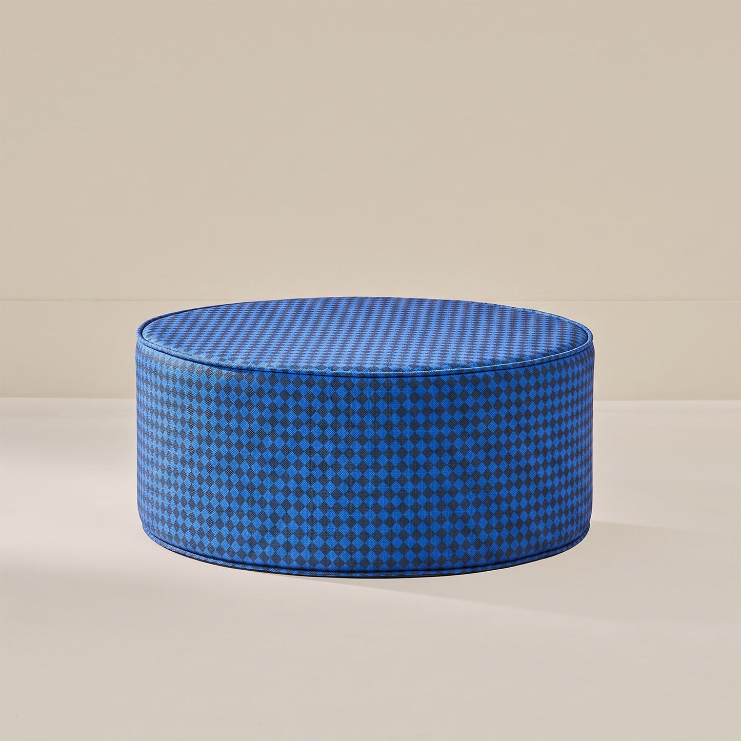 A round floor cushion or ottoman upholstered in a blue and black houndstooth pattern, set against a minimalist beige background.