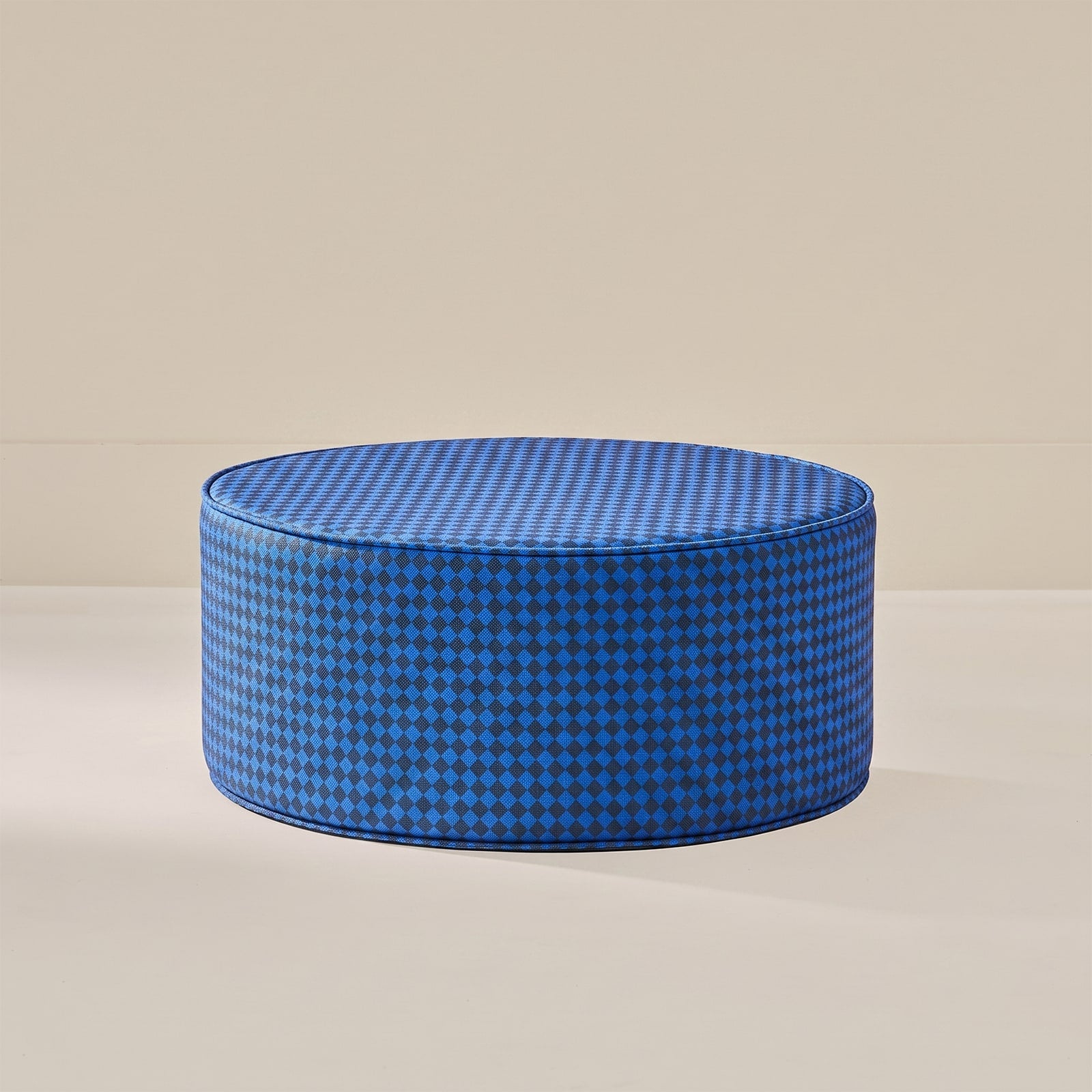 A round floor cushion or ottoman upholstered in a blue and black houndstooth pattern, set against a minimalist beige background.