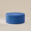 A round floor cushion or ottoman upholstered in a blue and black houndstooth pattern, set against a minimalist beige background.