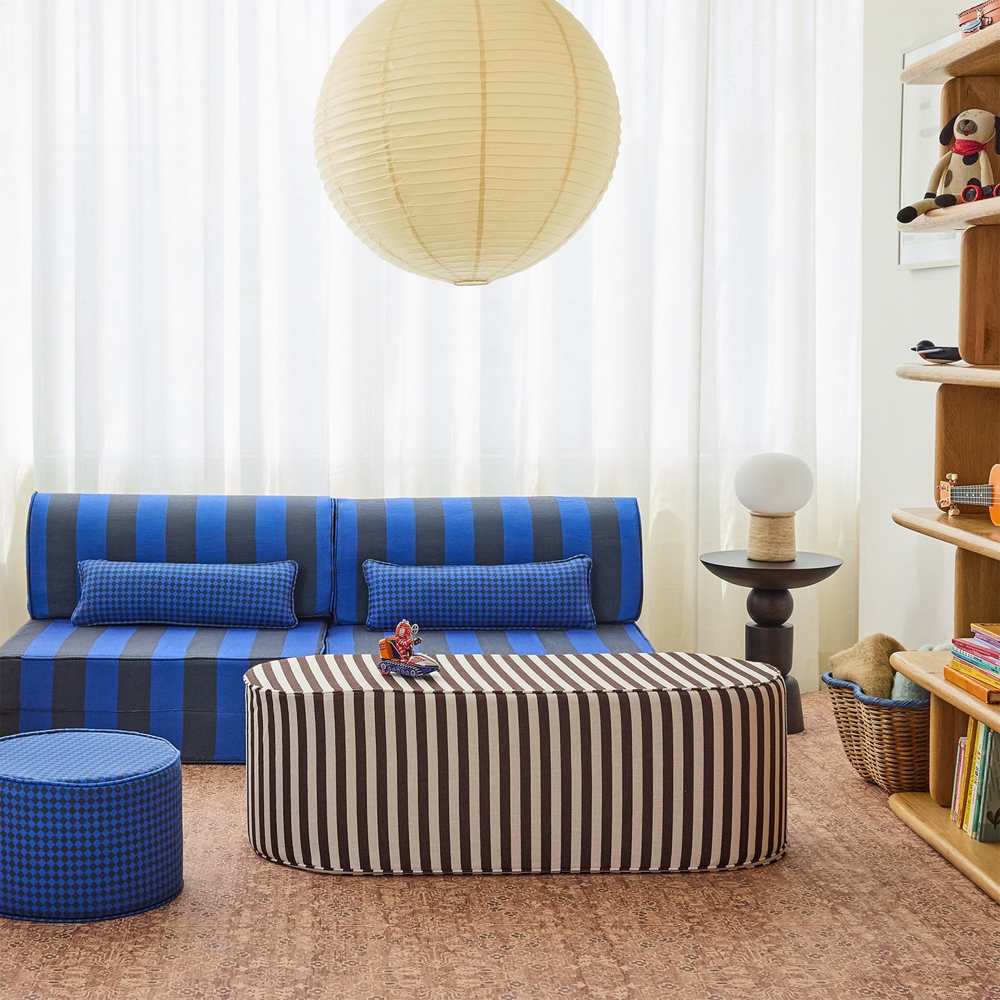 A styled living room scene featuring a blue and black striped sofa with matching pillows, a striped ottoman coffee table, a blue checkered pouf, and a large cream paper lantern hanging from the ceiling.