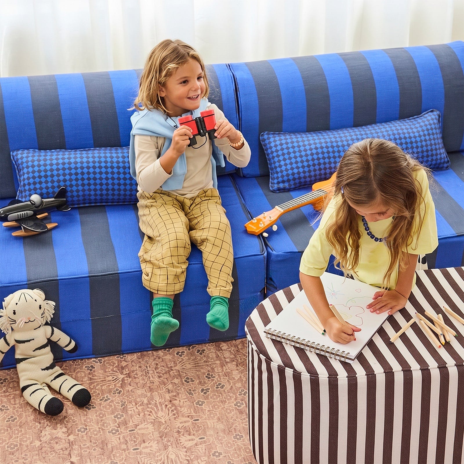 Two children playing on a blue and navy striped sofa in a brightly lit living room. One child sits holding a toy airplane while the other leans over a striped ottoman drawing on a notebook.