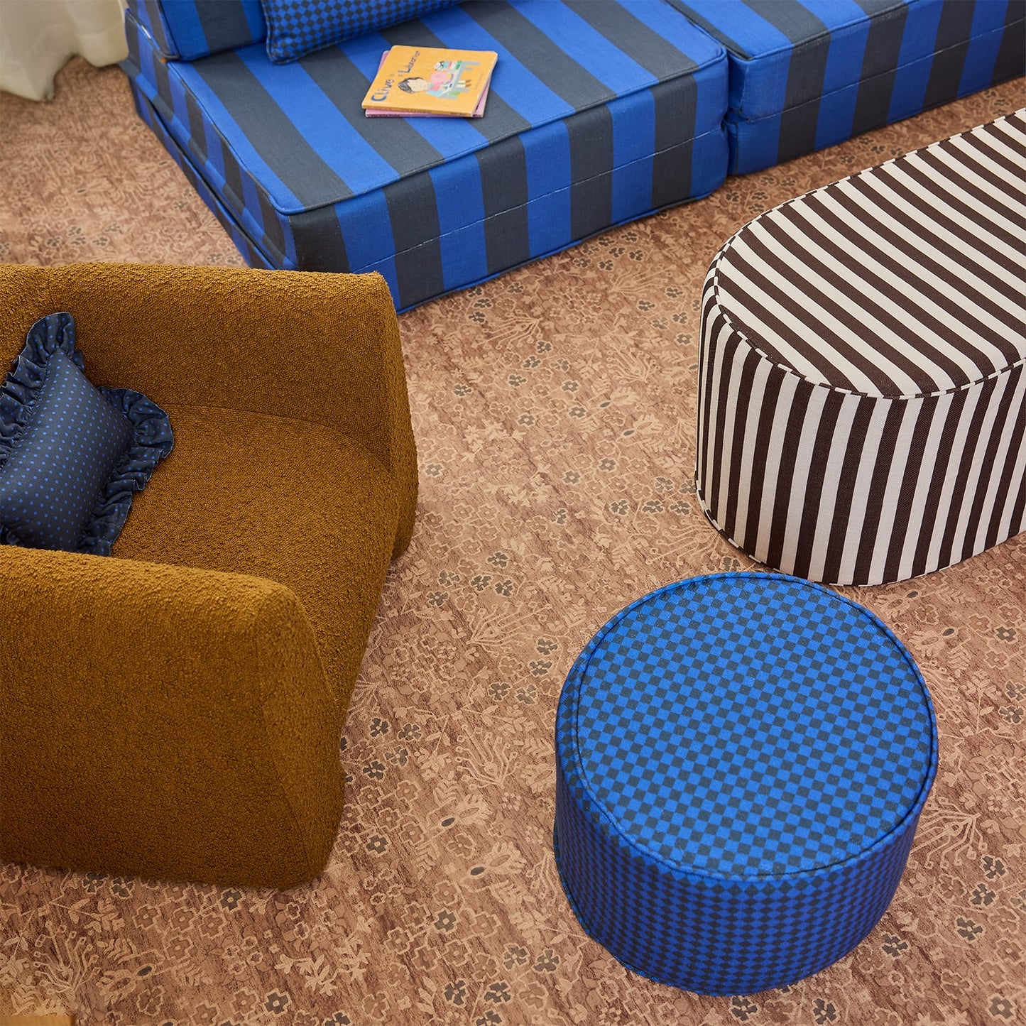 A styled interior scene showing multiple upholstered furniture pieces: a mustard-brown armchair with a blue ruffled pillow, a blue-and-gray striped sofa, a black-and-white striped ottoman, and a round blue-and-black checkered ottoman on a patterned beige rug.