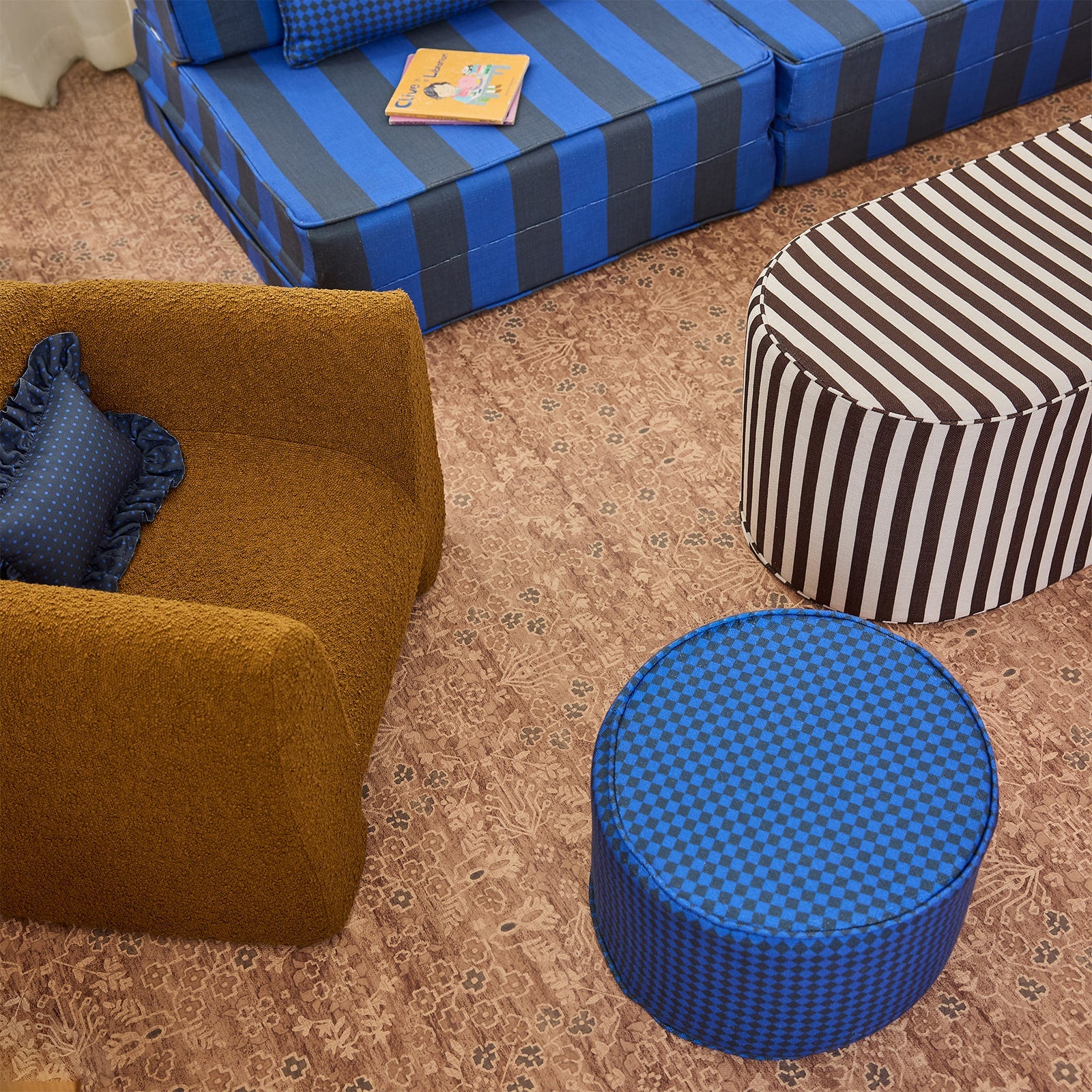 A styled interior scene showing multiple upholstered furniture pieces: a mustard-brown armchair with a blue ruffled pillow, a blue-and-gray striped sofa, a black-and-white striped ottoman, and a round blue-and-black checkered ottoman on a patterned beige rug.