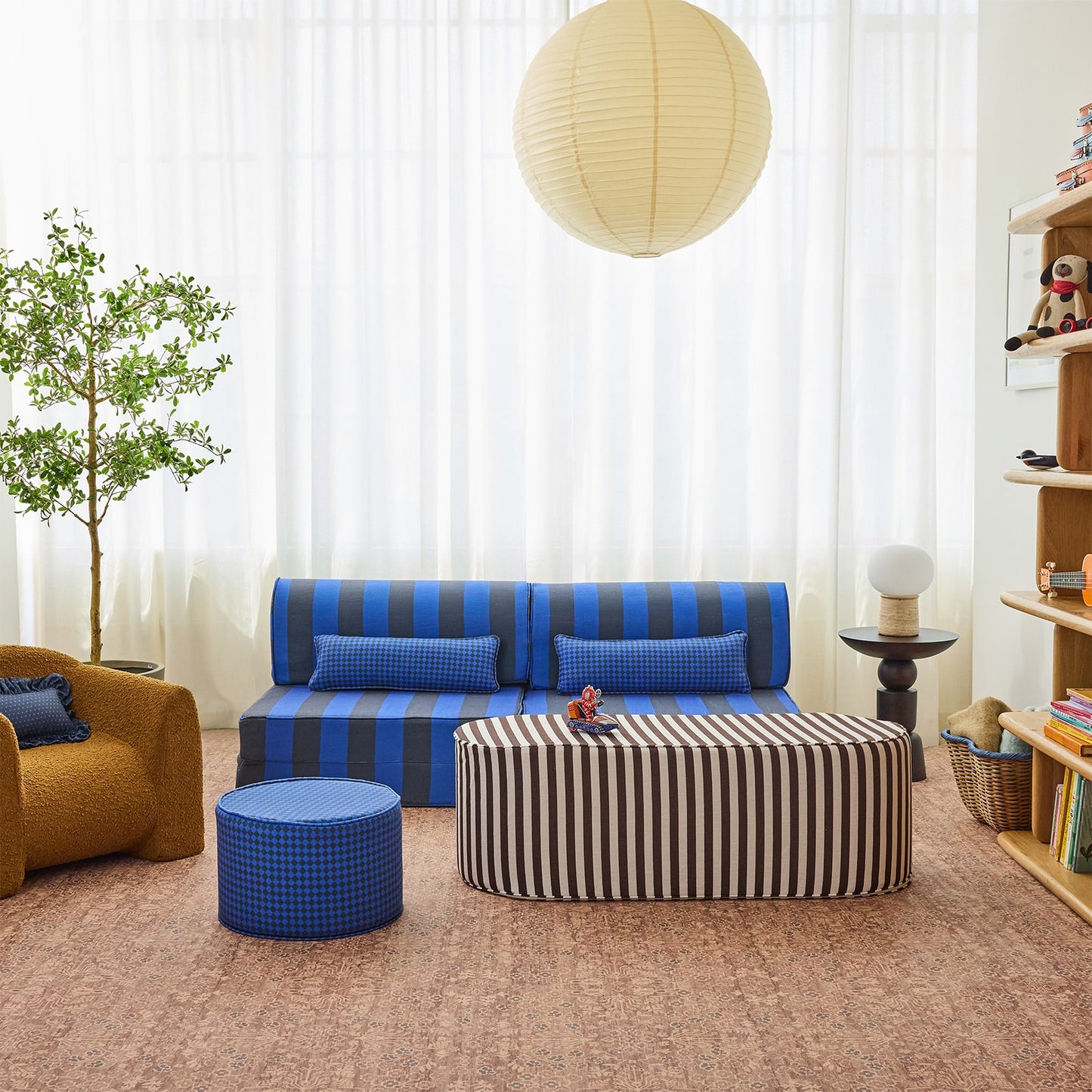 A styled children's playroom featuring a blue and black striped sofa, a matching blue ottoman, and a striped rectangular table, set against a background of sheer white curtains and a wooden bookshelf.