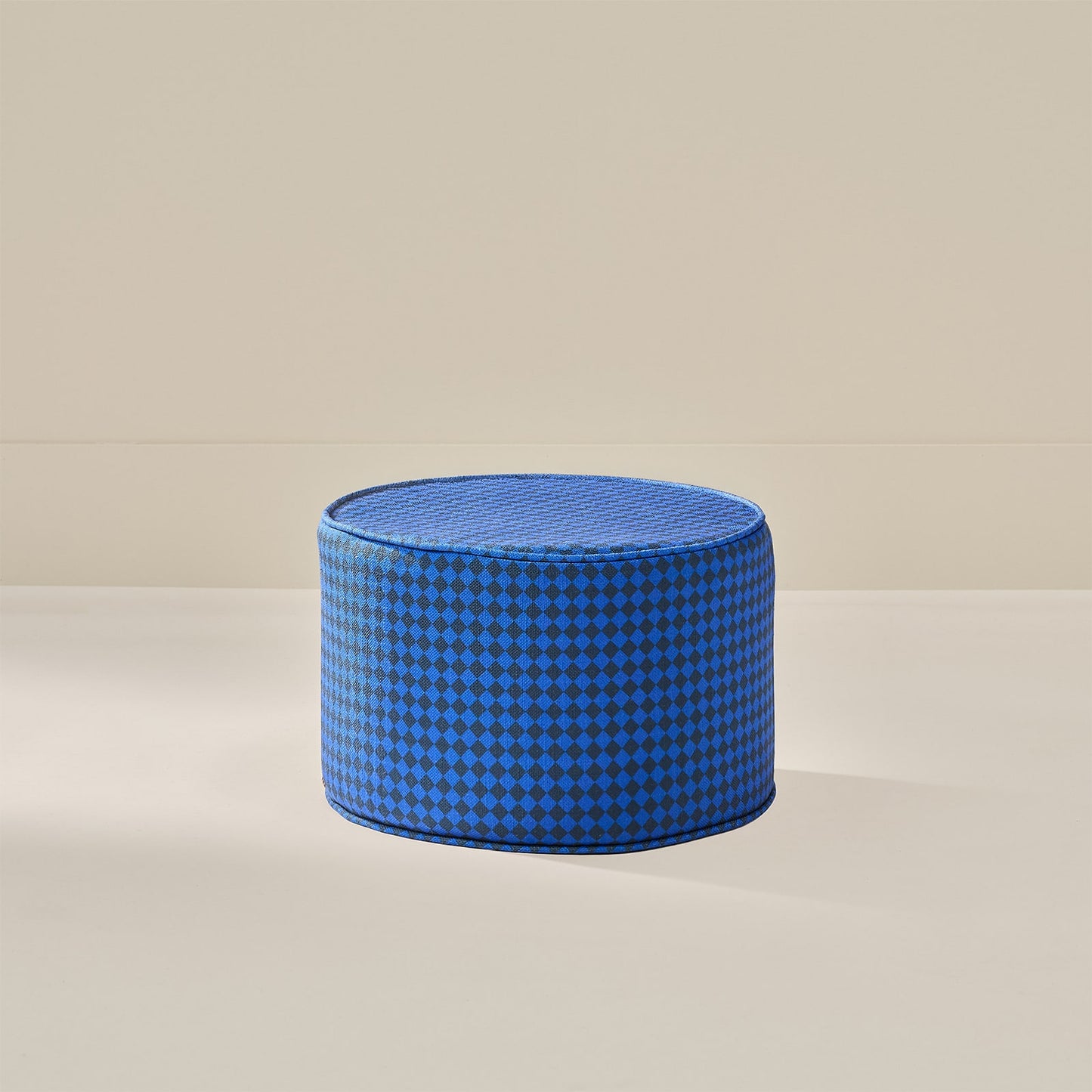 A round blue pouf ottoman with a small-scale blue and navy checkered pattern, set against a minimalist beige background.