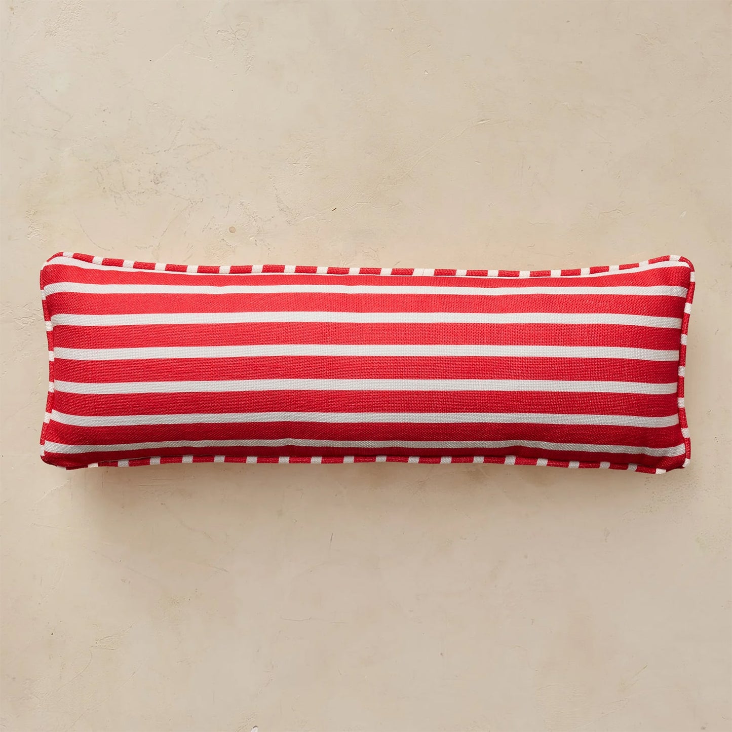 A rectangular lumbar pillow with horizontal red and white stripes, finished with a white dashed piped edge, resting on a textured beige surface.