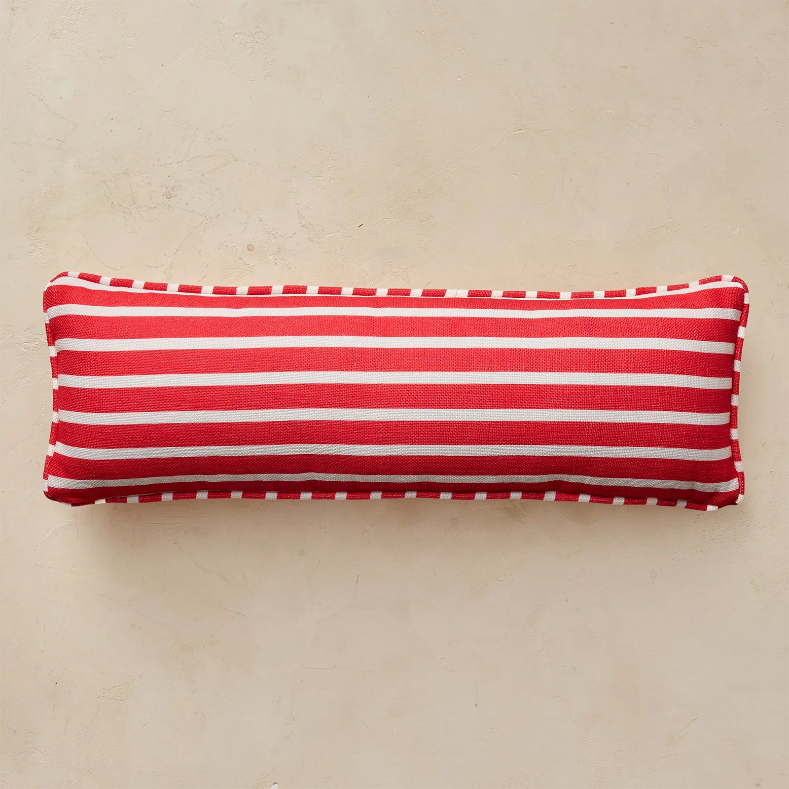 A rectangular lumbar pillow with horizontal red and white stripes, finished with a white dashed piped edge, resting on a textured beige surface.
