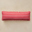A rectangular lumbar pillow with horizontal red and white stripes, finished with a white dashed piped edge, resting on a textured beige surface.