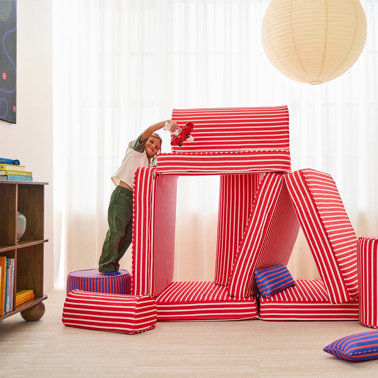 A child stands on an ottoman large red and white striped foam play castle in a bright room with white curtains, a hanging paper lantern, and a wooden bookshelf.