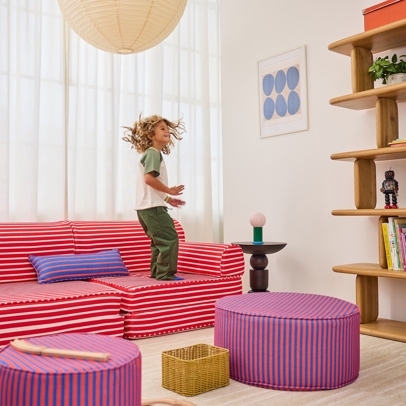 A child stands on a red-and-white striped sofa in a bright, modern room with pink-and-blue striped ottomans, a wooden bookshelf, and white curtains.