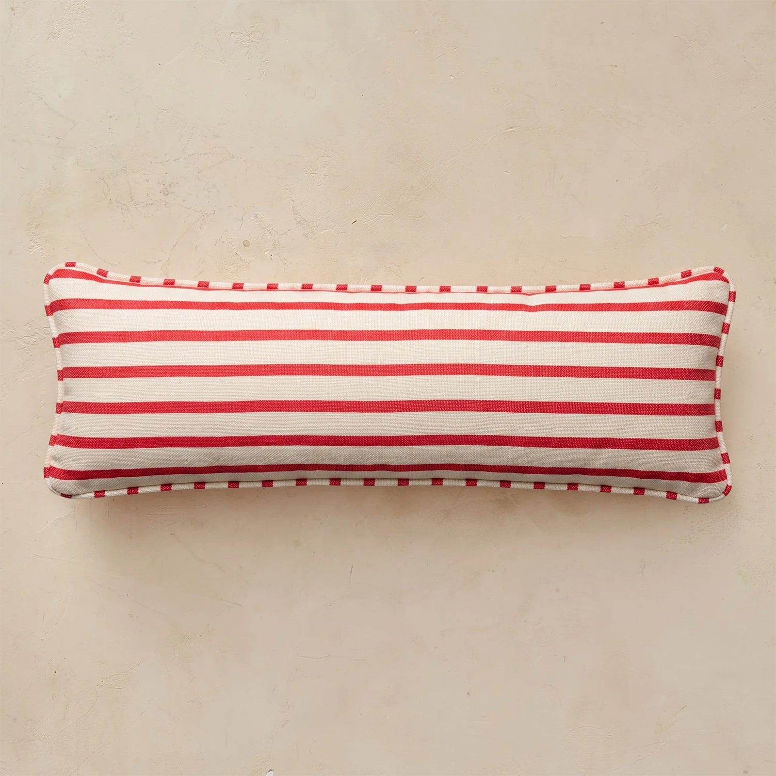 A rectangular lumbar pillow with horizontal red and white stripes and a red-and-white checkered piped edge, shown on a textured beige background.