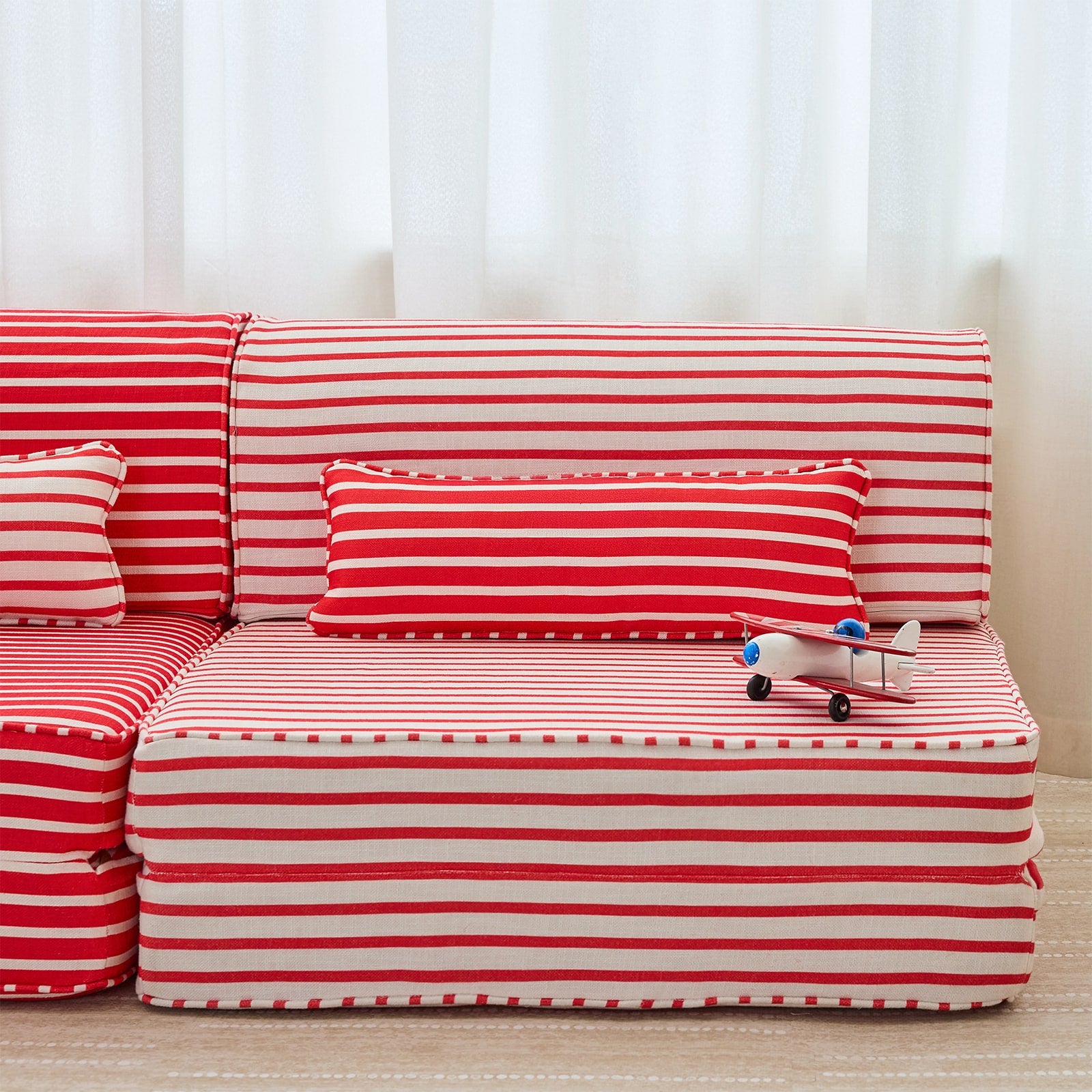 Red and white striped sofa cushions set arranged on a sofa, with a matching rectangular throw pillow and a small toy airplane resting on the seat cushion, against a soft white curtain background.