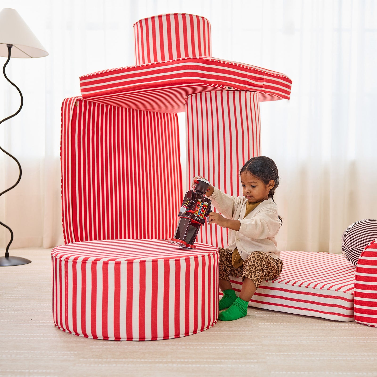 A child sits on a red and white striped play couch thats been stacked like a fort, playing with a toy.