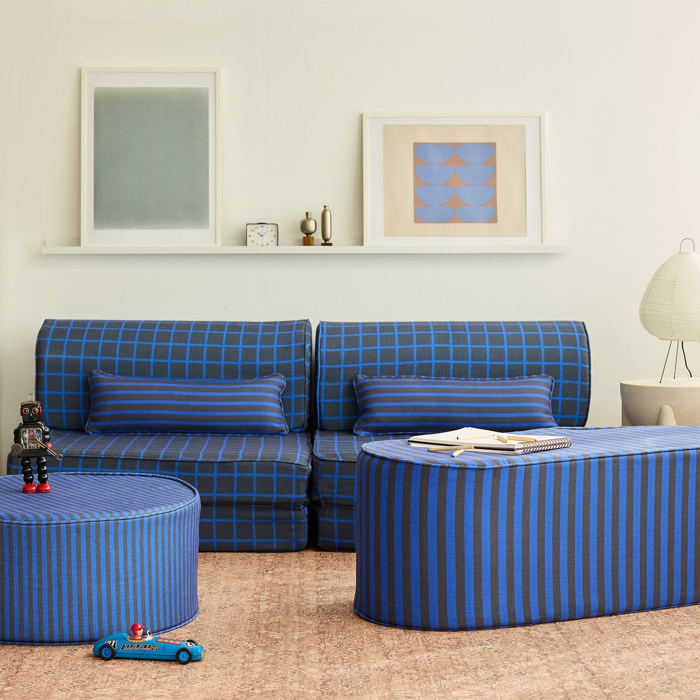 A styled children's living room featuring a blue and navy grid pattern modular sofa, a matching striped ottoman coffee table, and a striped cylindrical side table on a cork rug.