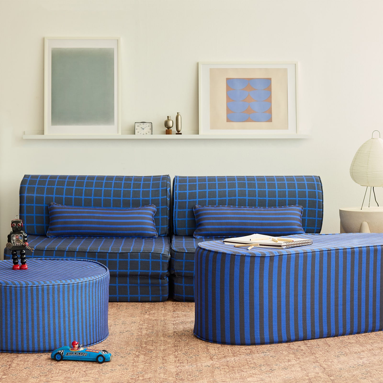 A styled children's living room featuring a blue and navy grid pattern modular sofa, a matching striped ottoman coffee table, and a striped cylindrical side table on a cork rug.