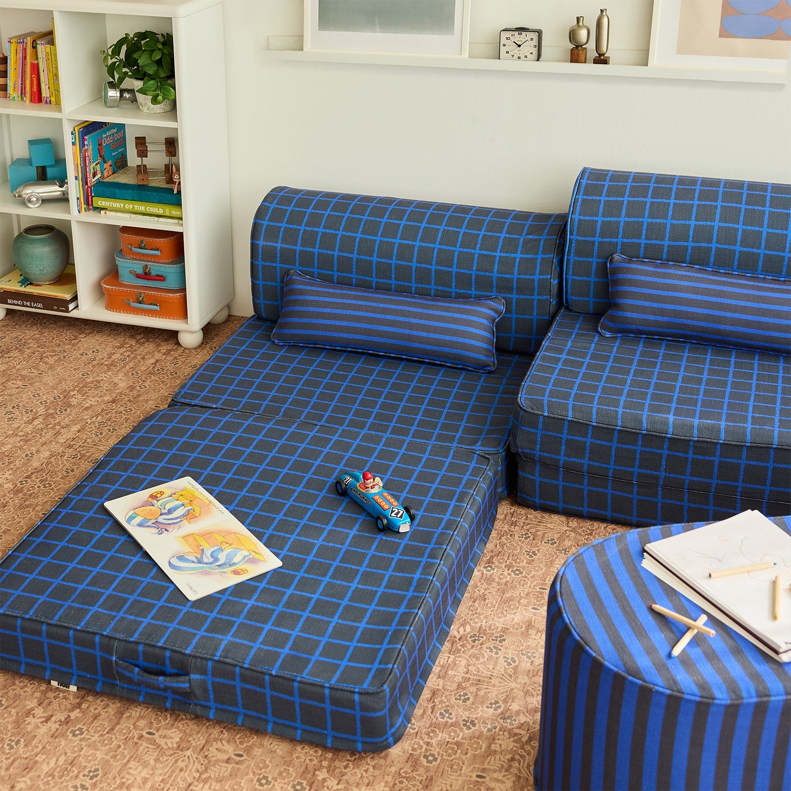 A blue and navy grid pattern modular play couch with matching pillows, styled in a child's room next to a white bookshelf and a matching striped ottoman.