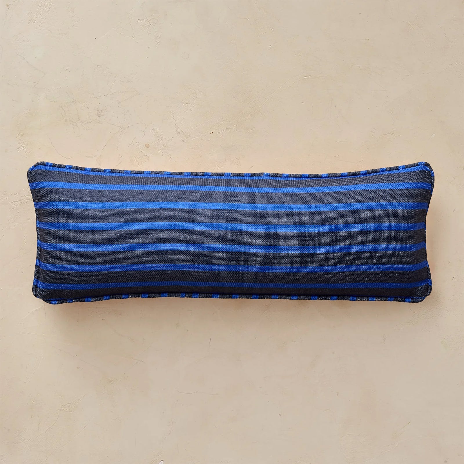A rectangular navy lumbar pillow with horizontal blue stripes, shown against a neutral beige textured background.