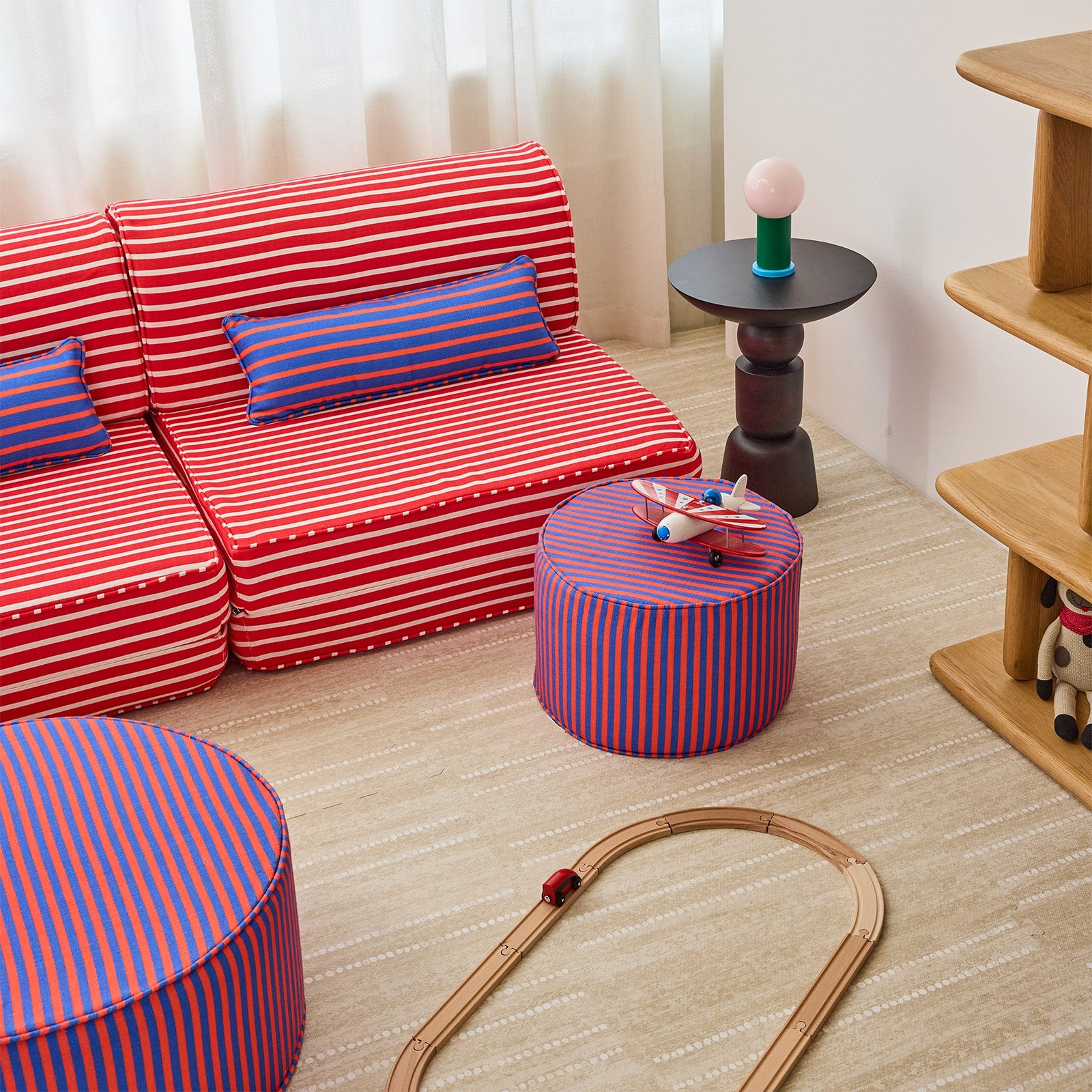 A children's room furniture set featuring a red and white striped sofa, matching striped pillows, a red and white striped ottoman, and a blue and red striped cylindrical stool, arranged on a light wood floor in a styled room.