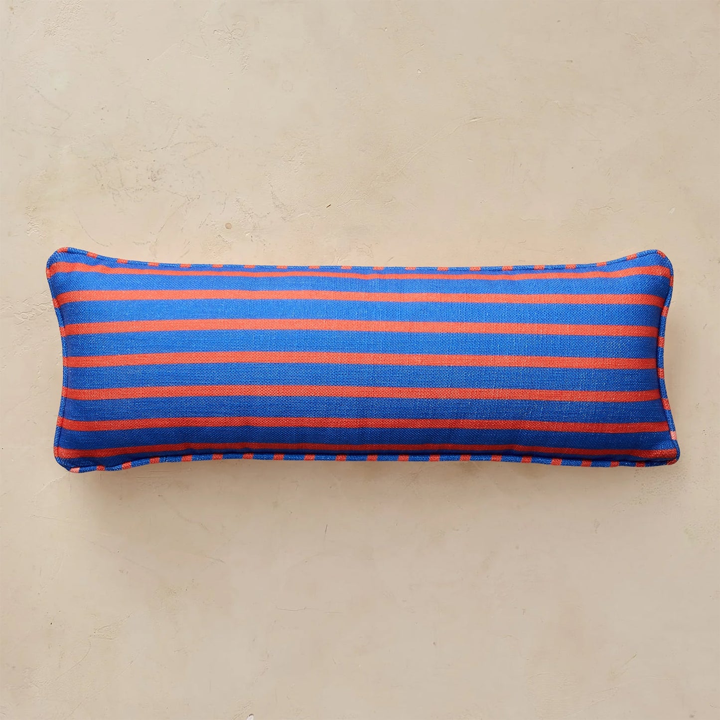 A rectangular lumbar pillow with horizontal stripes in royal blue and orange, resting on a textured beige surface.